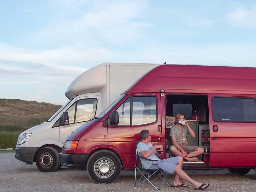 red campervan parked on the beach with two men drinking from cups